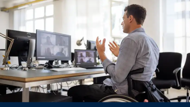 Image shows a man sitting at a desk in a wheelchair. He is gesticulating with his hands, as if in a meeting with someone on the computer in front of him. He is surrounded by similar desks and computers, as in an shared office environment.