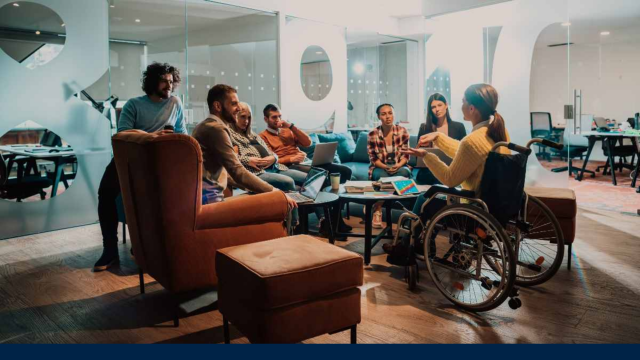 A group of young office workers are gathered in what appears to be a break-out area within an office environment. Some are seated on a large sofa and comfy armchairs, while another is perched on the arm of one of the sofas and one of the group is seated in a wheelchair and appears to be leading the meeting.