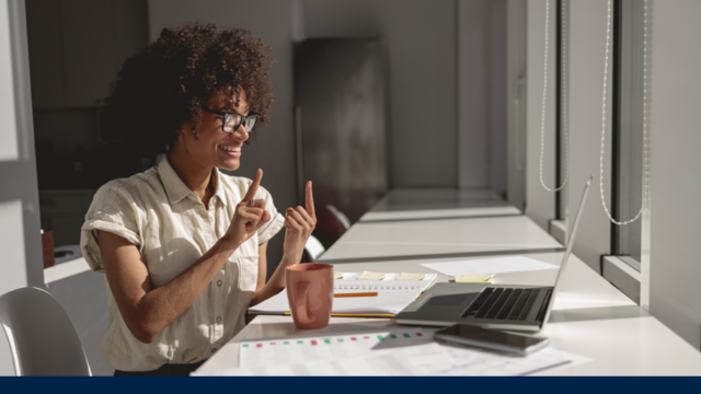 A young black female in an office setting smiles into a screen and appears to be having a conversation in sign language with someone on her laptop.