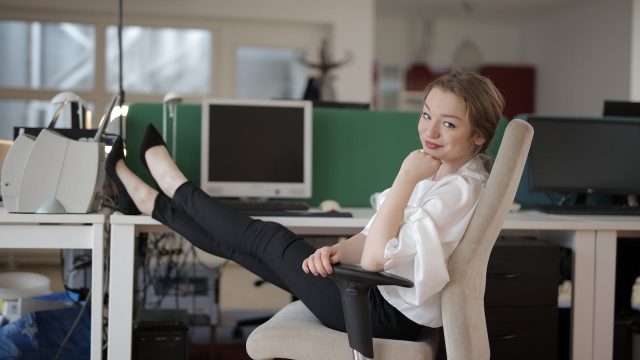 Image of a gen z female dressed in business wear and sitting back in a chair with her feet propped up in the desk in front of her. She's turned to the camera smiling.