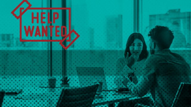 Two people sitting in an office setting and text to the left hand side reads "Help wanted" as if sellotaped to the image