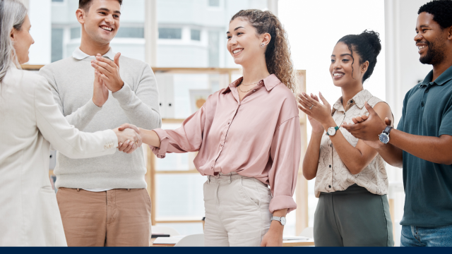 A group of young adults in business casual wear in an open office setting, one of the group is shaking hands with an older women in businesswear.