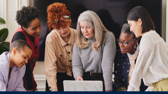 A group of diverse people in a professional setting gather around a laptop.
