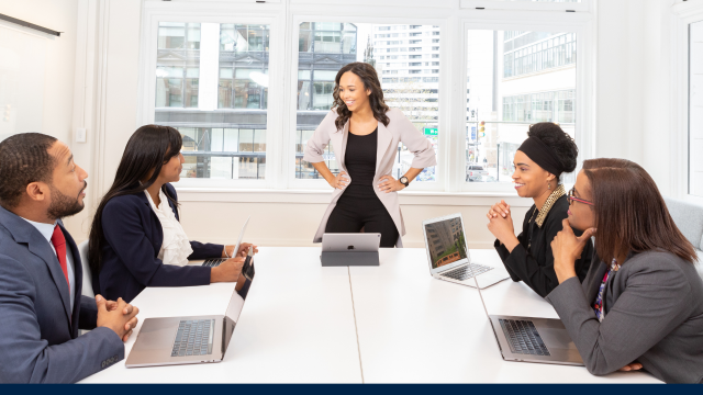 Group of people sit in a boardroom. A female is standing at the head of the table, talking to the rest.