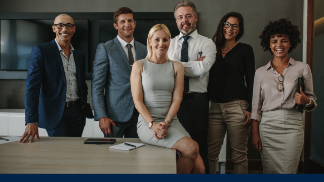 A diverse group of people in business/ business casual wear clothing. They appear to be in a large boardroom standing behind a large desk.