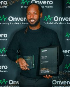 DEI Influencer of the Yea, Yohanes Scarlett, poses in front of the step and repeat banner with his award and certificate.