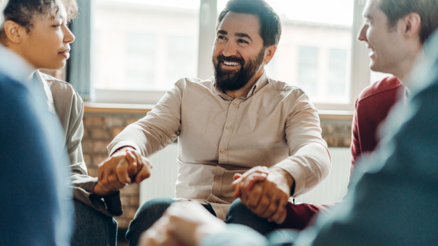 The image depicts a group of people seated in a circle indoors. In the centre, a man with a beard, wearing a beige shirt, smiles broadly. He holds hands with two other individuals, creating a sense of connection. On the left, a woman with curly hair and a white top looks at him attentively. To the right, another person is partly visible, engaged in conversation.