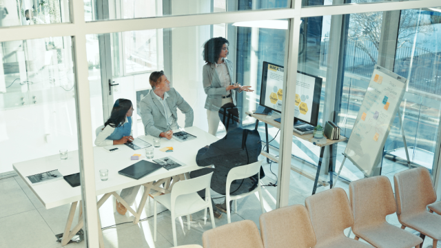 The image depicts a modern office meeting room with four diverse people engaged in a presentation. One person stands near a large display screen, gesturing as they speak. The screen shows a presentation slide with a title and graphical data.