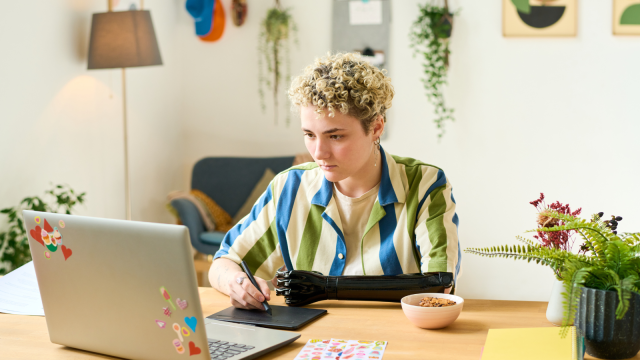 The image shows a person working at a wooden desk in a well-lit office. The individual is using a digital pen and tablet connected to an open laptop, which has colourful stickers on the back. The person's right arm appears to be a prosthetic.