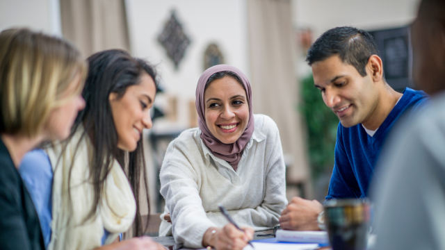The image depicts a group of three people engaged in conversation at a table. A woman in the centre, wearing a light purple hijab, smiles as she writes on a piece of paper. To her left, a woman is focused on the conversation. To her right, a man is also participating. In the top left corner, in white text, reads "Employee Network Hub." The bottom right corner displays the Onvero logo.