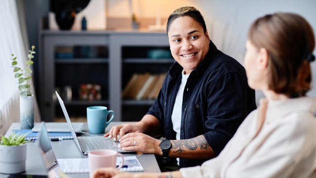The image shows two people sitting at a table in a bright, modern interior space. They are engaged in a friendly conversation, with one person smiling at the other. The person on the left, a masculine-presenting woman with a braided hairstyle, is wearing a black shirt and using a laptop.
