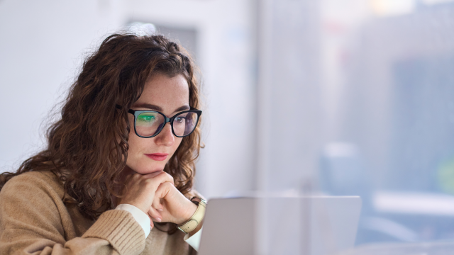 The image depicts a woman with long, curly brown hair and black-rimmed glasses, seated in front of a laptop. She is resting her chin on her hand, appearing focused on the screen. The background is softly blurred, contributing to the calm atmosphere, and features soft, diffused lighting.