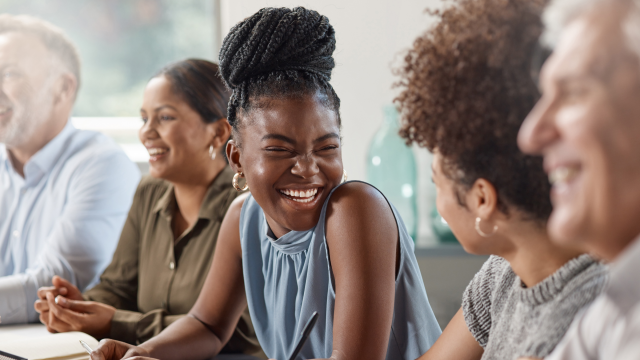 The image shows a group of five people sitting indoors, engaged in conversation and laughter. The centre person, a Black woman with braided hair styled in an updo, is smiling broadly, looking towards another person on her right. The atmosphere appears relaxed and joyful.